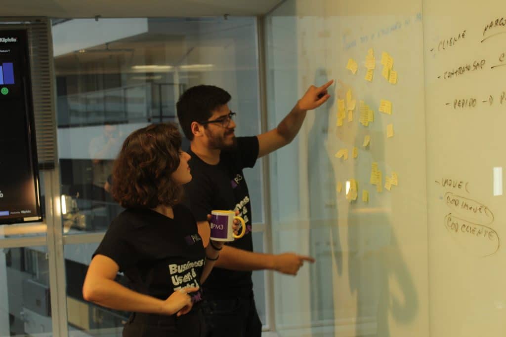 A imagem mostra Marcell Almeida e Natalia Arsand, profissionais da área de Gestão de Produtos, olhando para um quadro com post-its.