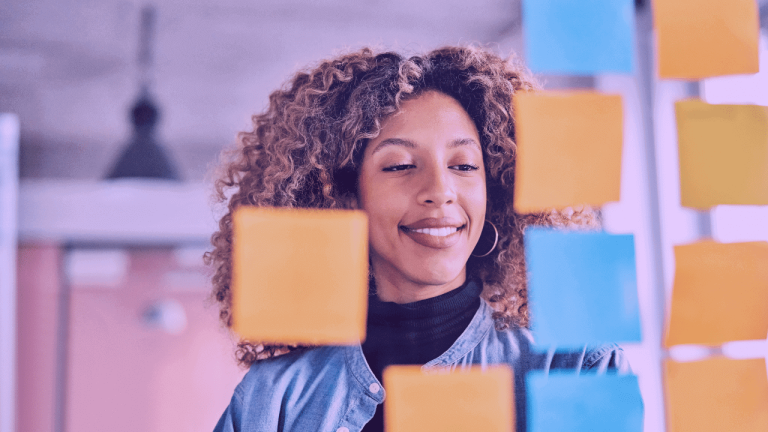 A imagem mostra uma mulher de frente para um quadro com post-its nas cores amarelo e azul. A foto sugere um processo de plane
