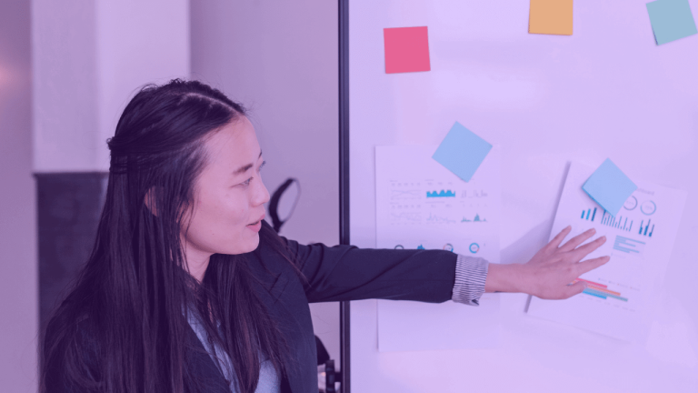 A imagem mostra uma mulher apontando para um quadro com post-its e gráficos de barra. A foto sugere uma apresentação de pitch