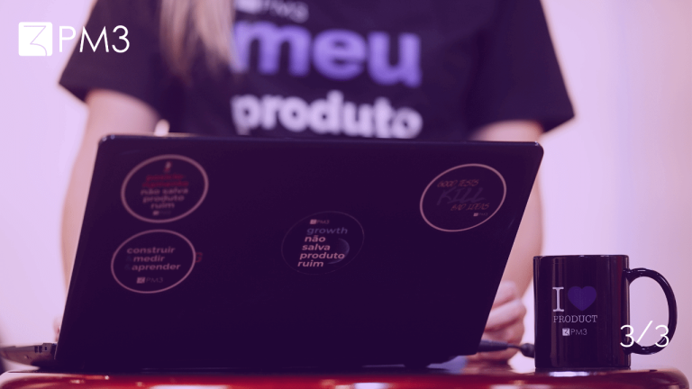 A imagem mostra um computador com adesivos da PM3 em primeiro plano, com a camisa de uma das instrutoras ao fundo